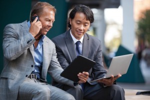 Two men looking at tablet and laptop while talking on cell phone